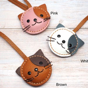 May include: Three leather cat-shaped keychains in pink, white, and brown. Each keychain has a brown leather strap and a stitched face with black button eyes and whiskers. The word "Pink", "White", and "Brown" are written below each keychain.