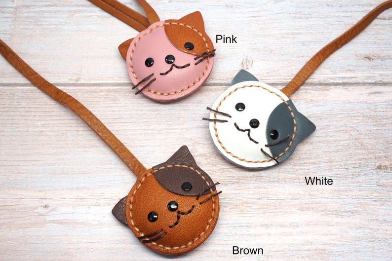 May include: Three leather cat-shaped keychains in pink, white, and brown. Each keychain has a brown leather strap and a stitched face with black button eyes and whiskers. The word "Pink", "White", and "Brown" are written below each keychain.