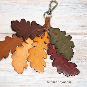 May include: A keychain featuring multiple leather oak leaf cutouts in shades of dark brown, tan, yellow, olive green, and deep red. The leaves are attached to a braided leather cord with a metal lobster clasp. The text Sunset Keychain is visible.