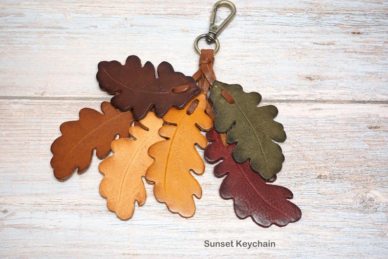 May include: A keychain featuring multiple leather oak leaf cutouts in shades of dark brown, tan, yellow, olive green, and deep red. The leaves are attached to a braided leather cord with a metal lobster clasp. The text Sunset Keychain is visible.