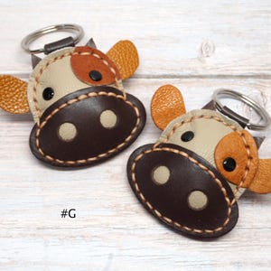 May include: Two cow-shaped keychains made of stitched leather in shades of cream, dark brown, and textured orange. Each keychain features a metal ring, black circular eyes, and a dark brown snout with two cream-colored nostrils. The text #G is visible.