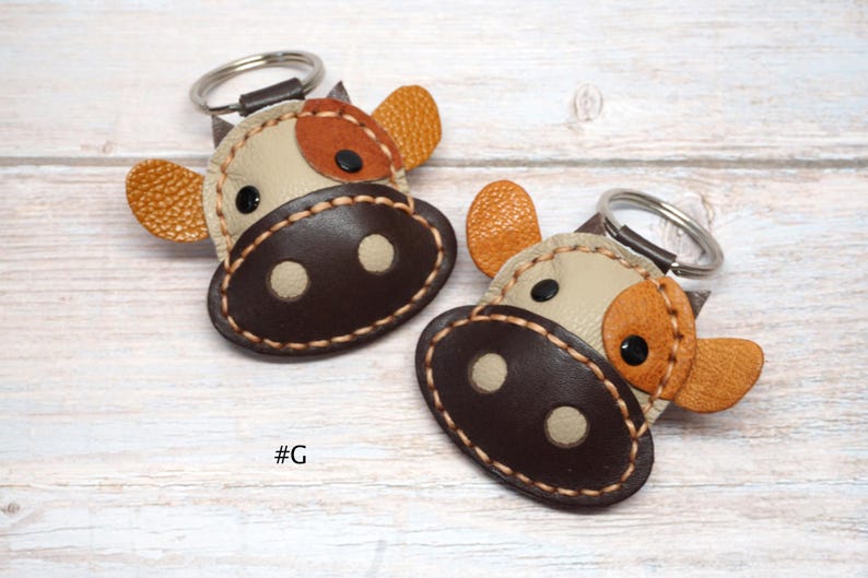 May include: Two cow-shaped keychains made of stitched leather in shades of cream, dark brown, and textured orange. Each keychain features a metal ring, black circular eyes, and a dark brown snout with two cream-colored nostrils. The text #G is visible.