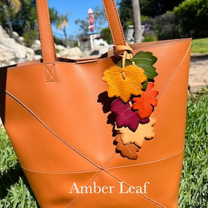 May include: A tan leather tote bag featuring two shoulder straps and a decorative cluster of multi-colored leather leaves in shades of green, yellow, orange, red, and beige attached to the handle. The text Amber Leaf is printed on the front.
