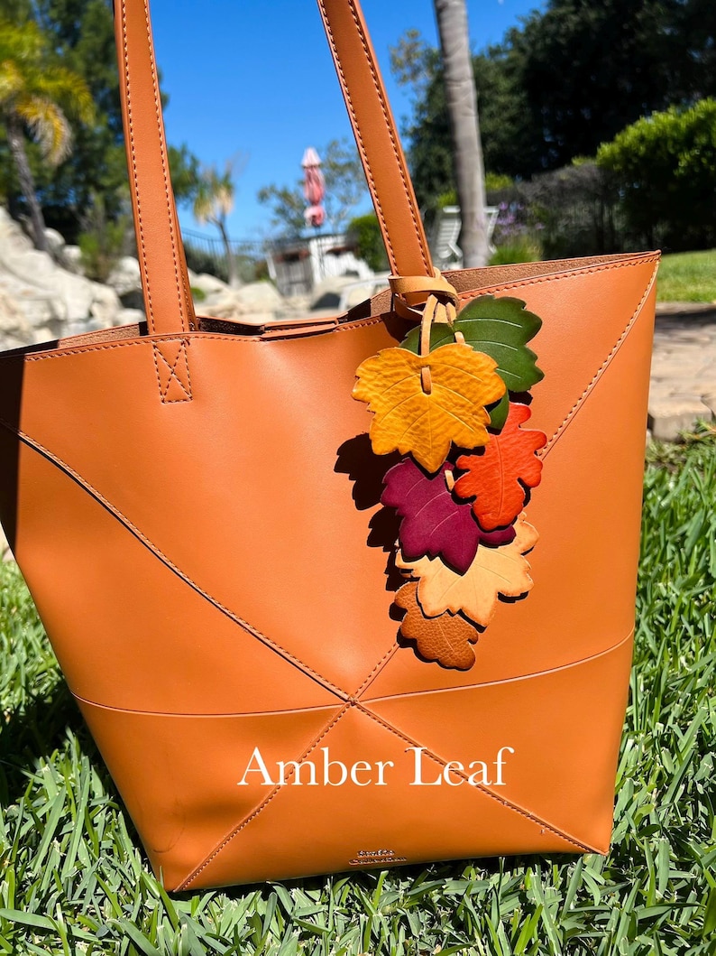 May include: A tan leather tote bag featuring two shoulder straps and a decorative cluster of multi-colored leather leaves in shades of green, yellow, orange, red, and beige attached to the handle. The text Amber Leaf is printed on the front.