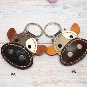 May include: Two leather cow-shaped keychains with metal rings. Keychain A features a dark brown textured snout, grey face, and brown ears. Keychain B features a dark brown snout, light grey face, and orange ears. Both have white eye patches.