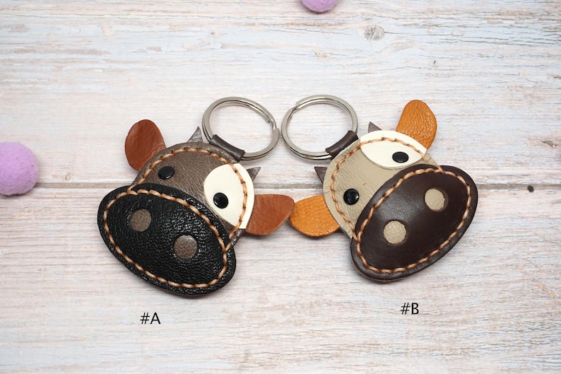 May include: Two leather cow-shaped keychains with metal rings. Keychain A features a dark brown textured snout, grey face, and brown ears. Keychain B features a dark brown snout, light grey face, and orange ears. Both have white eye patches.