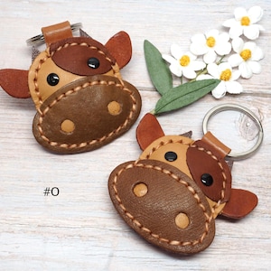 May include: Two cow-shaped keychains crafted from tan and dark brown leather with visible stitching, black circular eyes, and light tan nostrils. Each keychain features a metal ring attachment and the text #0 is printed near the left keychain.