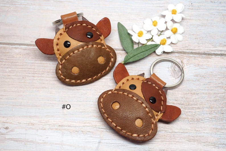 May include: Two cow-shaped keychains crafted from tan and dark brown leather with visible stitching, black circular eyes, and light tan nostrils. Each keychain features a metal ring attachment and the text #0 is printed near the left keychain.