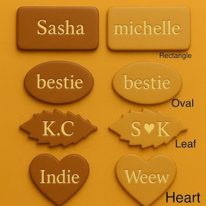 May include: Name tag options in tan and natural colors. Shapes include rectangle, oval, leaf, and heart. Each tag displays a name or initials, such as "Sasha," "Michelle," "Bestie," "K.C," "Indie," and "Weew."