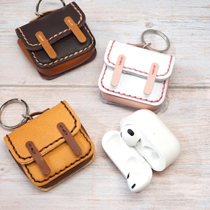 May include: Three leather cases for wireless earbuds, each designed like a miniature backpack with a metal keyring. One case is dark brown, one is white with pink stitching, and one is yellow with brown straps. A white charging case is open.