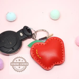 May include: A red leather apple-shaped keychain with a green leaf and yellow stitching. The keychain is attached to a car key fob. The words "GENUINE LEATHER" are stamped on the leather.