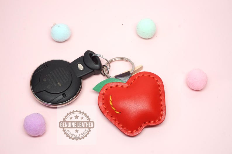 May include: A red leather apple-shaped keychain with a green leaf and yellow stitching. The keychain is attached to a car key fob. The words "GENUINE LEATHER" are stamped on the leather.