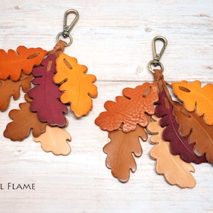 May include: Two keychains featuring clusters of die-cut leather oak leaves in shades of orange, yellow, brown, and deep burgundy. Each keychain is attached to a brass-toned metal lobster clasp. The text FALL FLAME is printed in the bottom left corner.