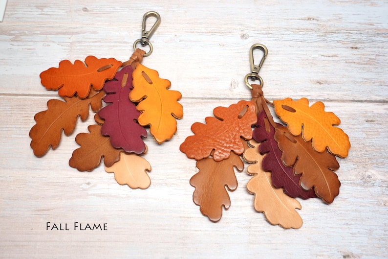 May include: Two keychains featuring clusters of die-cut leather oak leaves in shades of orange, yellow, brown, and deep burgundy. Each keychain is attached to a brass-toned metal lobster clasp. The text FALL FLAME is printed in the bottom left corner.