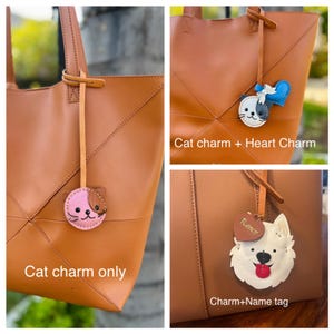 May include: A brown leather tote bag with a cat charm, a cat and heart charm, and a charm with a dog design. The cat charms are pink and black and white. The dog charm has a name tag.