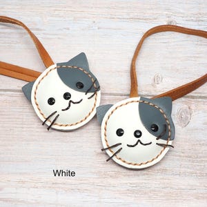 May include: Two white and gray cat-shaped leather keychains with brown leather straps. The cat faces have black eyes, whiskers, and a stitched outline. The word "White" is printed at the bottom.