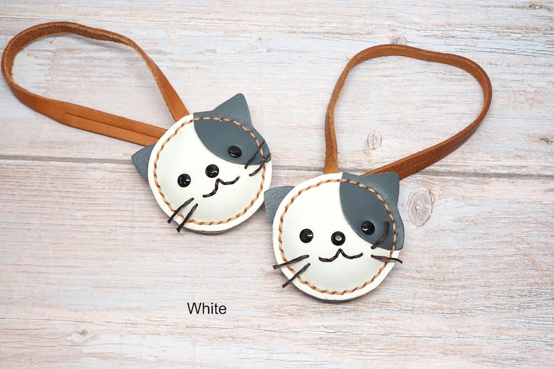 May include: Two white and gray cat-shaped leather keychains with brown leather straps. The cat faces have black eyes, whiskers, and a stitched outline. The word "White" is printed at the bottom.