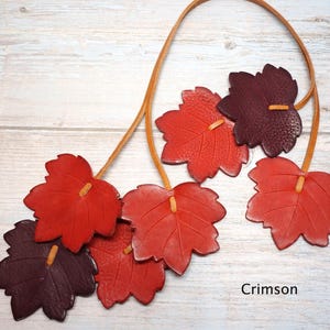 May include: A necklace featuring multiple leaf-shaped pendants in shades of red and dark burgundy, strung on a thin tan leather cord. The leaves have visible veins and textured surfaces. The word Crimson is printed in the bottom right corner.