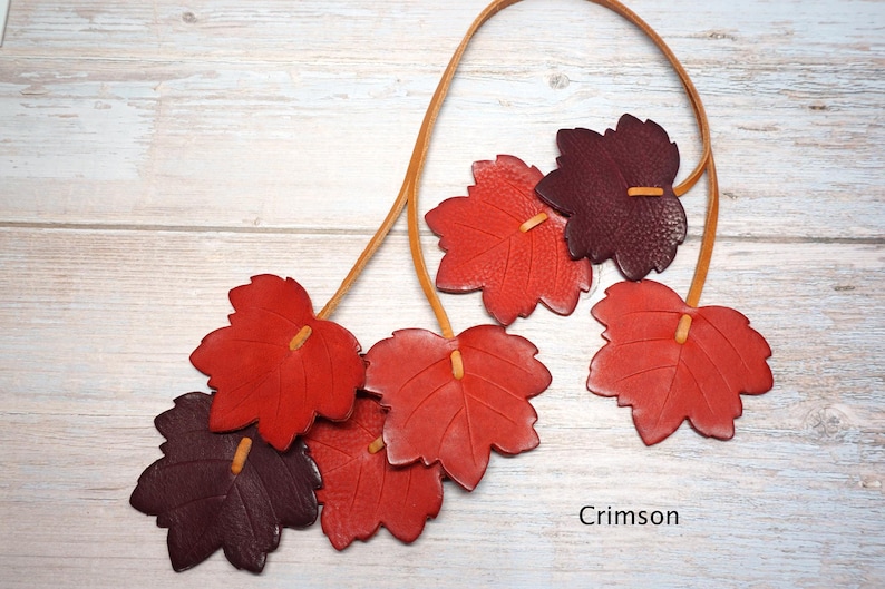 May include: A necklace featuring multiple leaf-shaped pendants in shades of red and dark burgundy, strung on a thin tan leather cord. The leaves have visible veins and textured surfaces. The word Crimson is printed in the bottom right corner.