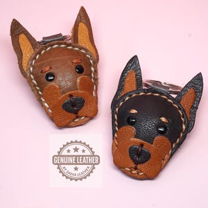 May include: Two dog-shaped keychains crafted from textured leather. One is brown with tan accents, and the other is black with tan accents. Both feature stitched edges, black bead eyes, and a metal ring. A logo reads GENUINE LEATHER BY SASHA LEATHER.