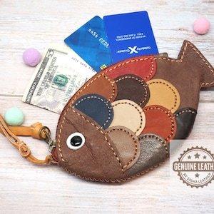 May include: A fish-shaped leather coin purse featuring multicolored circular patches in brown, tan, red, and blue, with a zipper closure, a wrist strap, and a white eye. The purse contains a five-dollar bill and two credit cards. A logo reads GENUINE LEATHER.