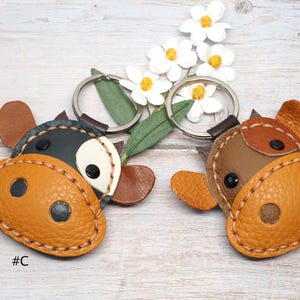 May include: Two leather cow-shaped keychains labeled #C and #D. The keychain labeled #C is black and white with a tan snout. The keychain labeled #D is brown and tan with a tan snout. Both feature metal rings and stitched edges.