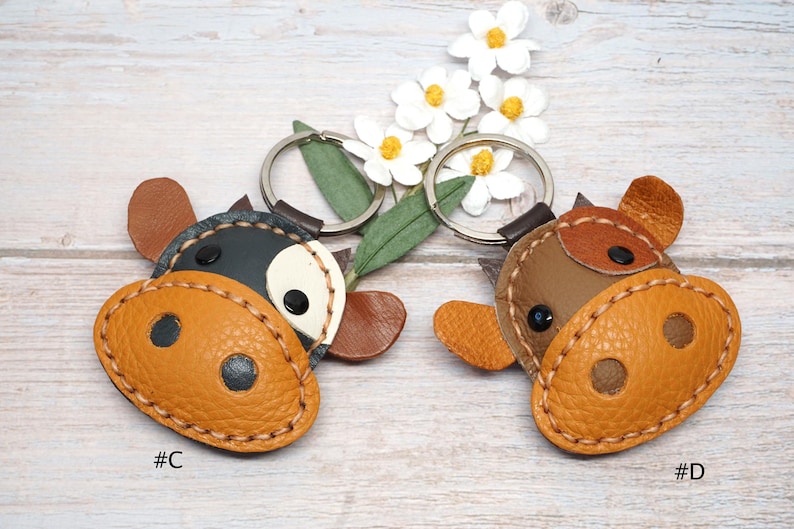 May include: Two leather cow-shaped keychains labeled #C and #D. The keychain labeled #C is black and white with a tan snout. The keychain labeled #D is brown and tan with a tan snout. Both feature metal rings and stitched edges.