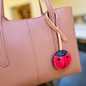 May include: A dusty rose handbag with two vertical stitched seams and two top handles. A circular leather charm shaped like a ladybird with a red body, black head, and black spots hangs from a light tan leather cord attached to the bag handle.