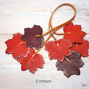 May include: A decorative hanging ornament featuring six leather maple leaves in shades of red and dark burgundy, attached to a thin tan leather cord. The leaves are textured with visible veins and the word Crimson is printed at the bottom.