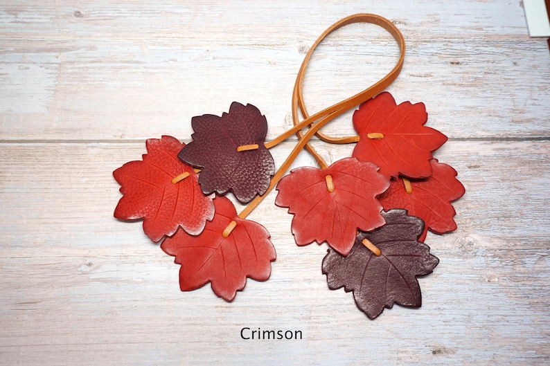 May include: A decorative hanging ornament featuring six leather maple leaves in shades of red and dark burgundy, attached to a thin tan leather cord. The leaves are textured with visible veins and the word Crimson is printed at the bottom.