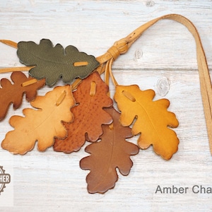 May include: A leather charm featuring six oak leaf cutouts in shades of olive green, tan, orange, and brown, attached to a long leather strap. The charm includes a circular logo with the text GENUINE LEATHER BY SASHA LEATHER and the text Amber Charm.