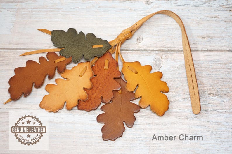 May include: A leather charm featuring six oak leaf cutouts in shades of olive green, tan, orange, and brown, attached to a long leather strap. The charm includes a circular logo with the text GENUINE LEATHER BY SASHA LEATHER and the text Amber Charm.