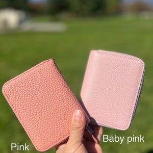 May include: Two pink leather wallets are displayed, one in a darker pink and the other in a lighter baby pink. Both wallets have a zip-around closure and a textured surface. The wallets are held up against a green background.