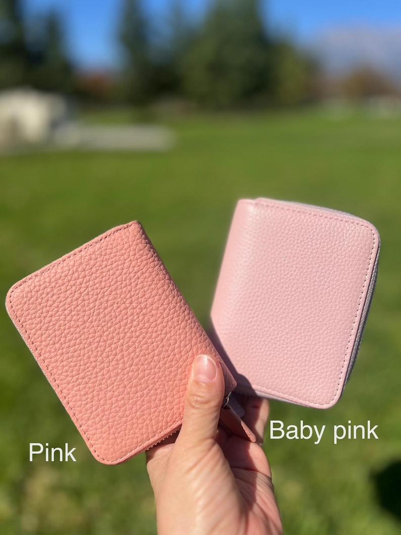 May include: Two pink leather wallets are displayed, one in a darker pink and the other in a lighter baby pink. Both wallets have a zip-around closure and a textured surface. The wallets are held up against a green background.