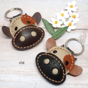 May include: Two cow-shaped keychains made of stitched leather with metal rings. One features a dark brown snout and beige face with brown patches, while the other has a black snout and beige face with tan patches. The text #M is visible.