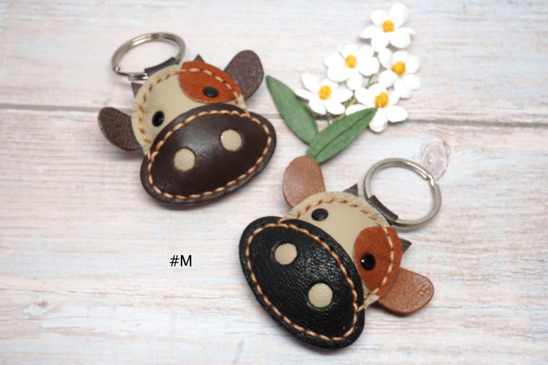May include: Two cow-shaped keychains made of stitched leather with metal rings. One features a dark brown snout and beige face with brown patches, while the other has a black snout and beige face with tan patches. The text #M is visible.