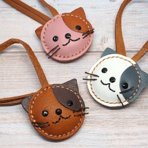 May include: Three handmade leather cat-shaped keychains in pink, brown, and white with brown leather straps. Each keychain features stitched details, black button eyes, and embroidered whiskers. The keychains are arranged on a light wood surface.