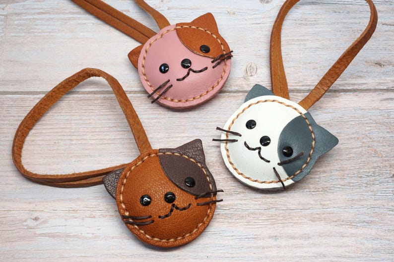 May include: Three handmade leather cat-shaped keychains in pink, brown, and white with brown leather straps. Each keychain features stitched details, black button eyes, and embroidered whiskers. The keychains are arranged on a light wood surface.