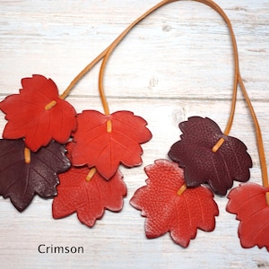May include: A necklace featuring multiple leather leaf-shaped pendants in shades of red and dark burgundy, suspended from thin tan leather cords. The leaves are textured and arranged in clusters, with the word Crimson printed at the bottom.