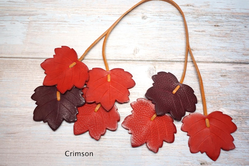 May include: A necklace featuring multiple leather leaf-shaped pendants in shades of red and dark burgundy, suspended from thin tan leather cords. The leaves are textured and arranged in clusters, with the word Crimson printed at the bottom.