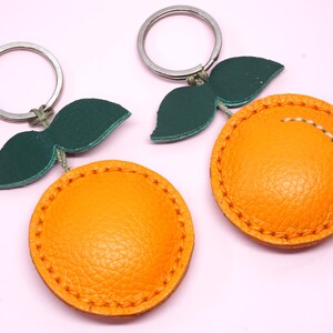 May include: Two orange-shaped keychains with green leaf accents and silver key rings. The orange portions are a textured, bright orange with stitched detailing. The leaves are a dark green. The background is a light pink.