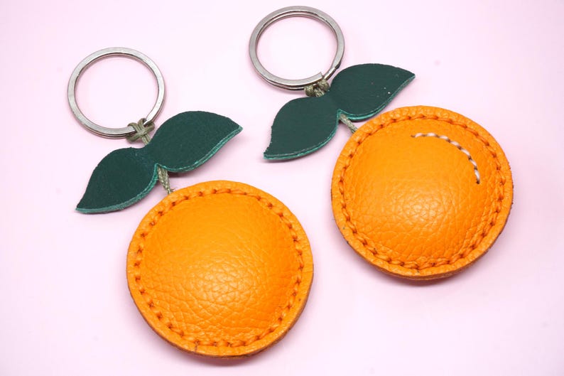 May include: Two orange-shaped keychains with green leaf accents and silver key rings. The orange portions are a textured, bright orange with stitched detailing. The leaves are a dark green. The background is a light pink.