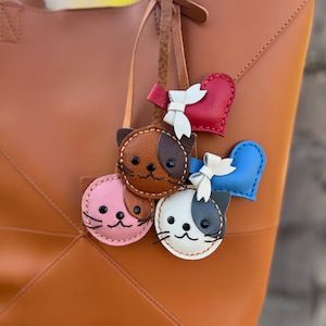 May include: A tan leather tote bag with a geometric design. Attached are several leather cat-shaped keychains in pink, brown, and white, and heart-shaped charms in red and blue. Each charm has a white bow.