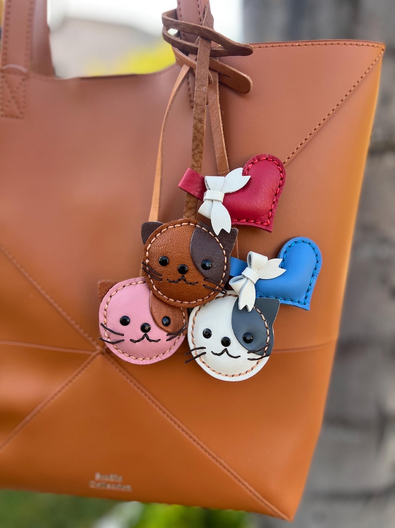 May include: A tan leather tote bag with a geometric design. Attached are several leather cat-shaped keychains in pink, brown, and white, and heart-shaped charms in red and blue. Each charm has a white bow.