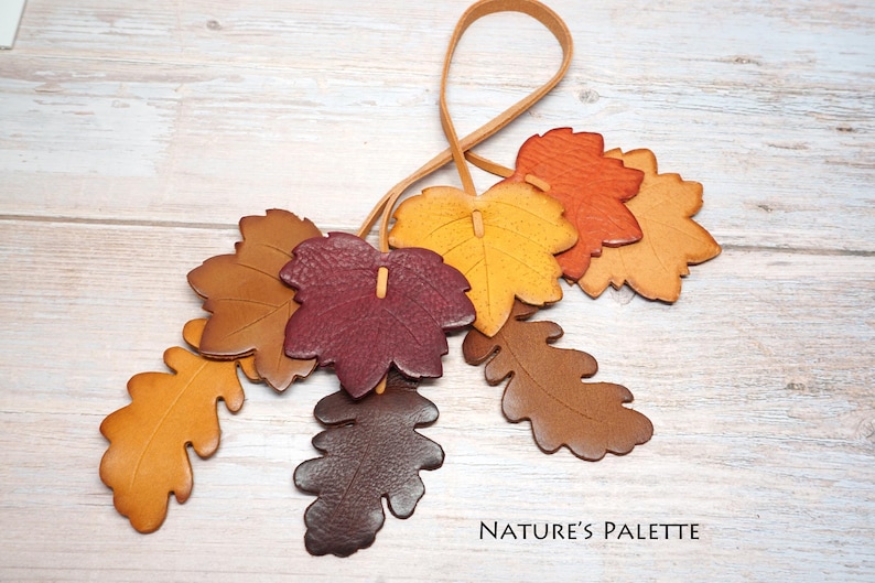 May include: A decorative hanging ornament featuring multiple leather leaves in shades of brown, orange, yellow, and deep burgundy, attached to a thin leather cord. The text "NATURE'S PALETTE" is printed in the bottom right corner of the image.
