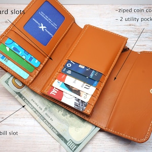 May include: A tan leather wallet featuring 9 credit card slots, a full-length bill slot, zipped coin compartments, and 2 utility pockets. The wallet is open, displaying various cards, a Celebrity Cruises ID, and a 100 dollar bill inside the bill slot.