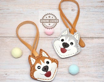 May include: Two leather dog-shaped keychains with tan leather loops. One keychain is brown and white, the other is gray and white. Both feature black noses, red tongues, and white stitching. A logo reads GENUINE LEATHER BY SASHA LEATHER.