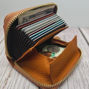 May include: Open, tan leather wallet with a gold zipper, filled with cards, cash, and coins. The wallet is square-shaped and has a textured surface. The interior features multiple card slots and a coin compartment.