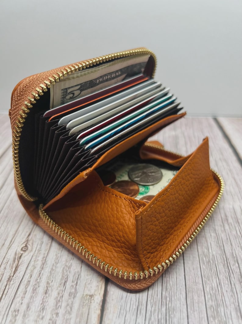 May include: Open, tan leather wallet with a gold zipper, filled with cards, cash, and coins. The wallet is square-shaped and has a textured surface. The interior features multiple card slots and a coin compartment.