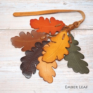 May include: A leather keychain featuring a cluster of six oak leaf-shaped cutouts in shades of orange, brown, yellow, and dark green. The leaves are attached to a thin leather strap with a loop. The text "EMBER LEAF" is printed in the corner.